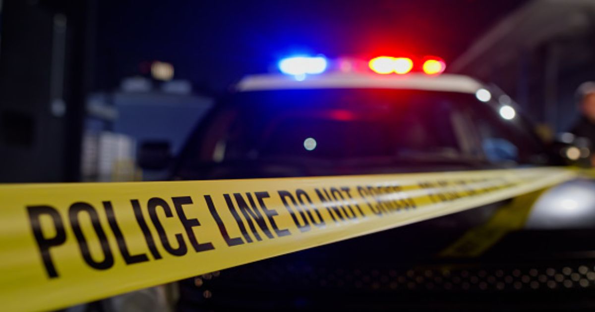 A crime scene is visible with the police lights flashing on the top of a car behind police tape.