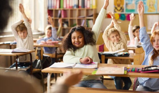 Children in a classroom sit with their hands raised toward the teacher.