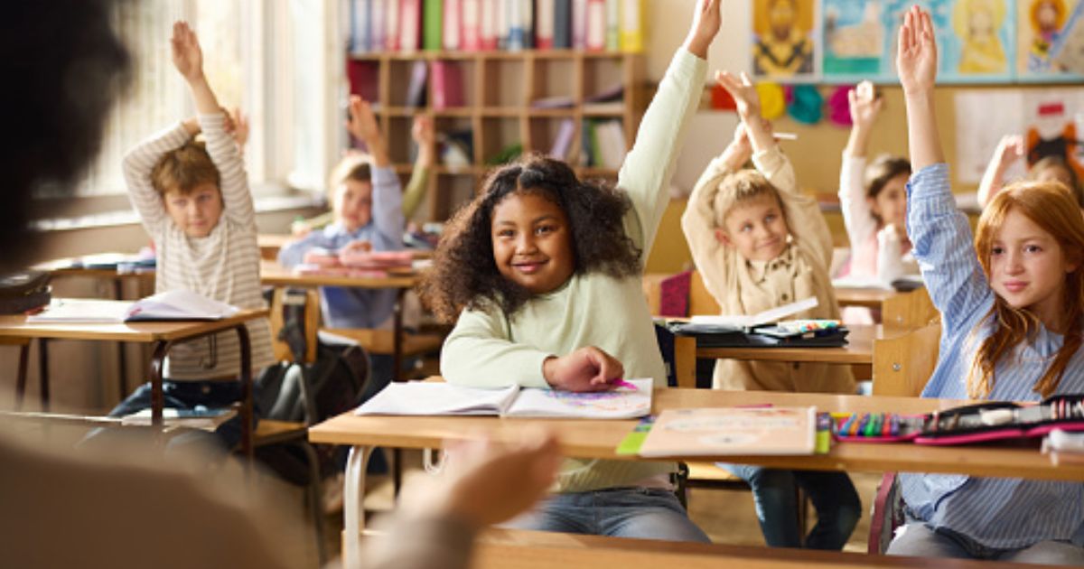 Children in a classroom sit with their hands raised toward the teacher.