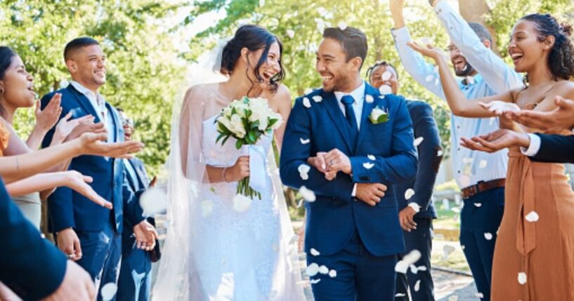 A couple gets married outdoors as attendees throw flower petals on them.
