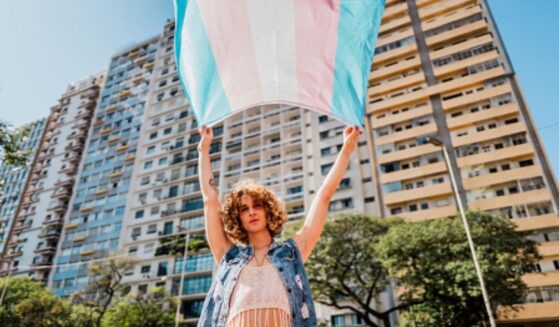A trans person waves a transgender flag near a large building.