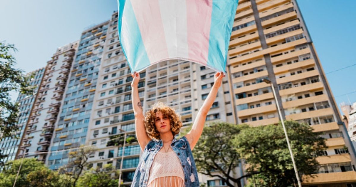 A trans person waves a transgender flag near a large building.