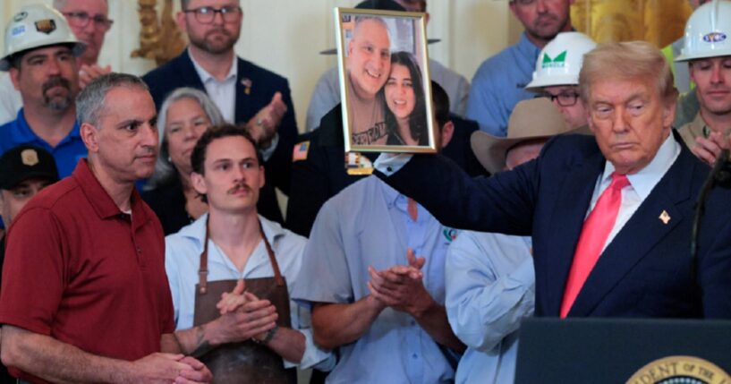 Joe Abraham, left, in red shirt, watches as President Donald Trump holds up a photograph of Abraham and his daughter, Katie Braham, during an event in the East Room of the White House in June 2025.