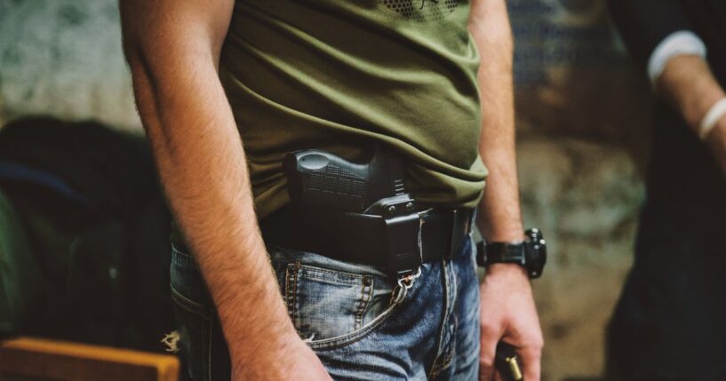 A person wearing jeans and a green shirt with a handgun holstered at their waist.