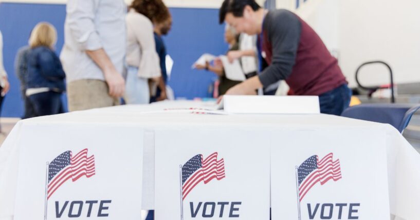 Ballots and American flags displayed on a table at a voting station, people are in the background.