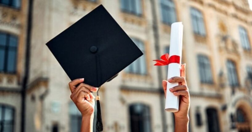 A student raises their degree and their graduation cap at a college campus.