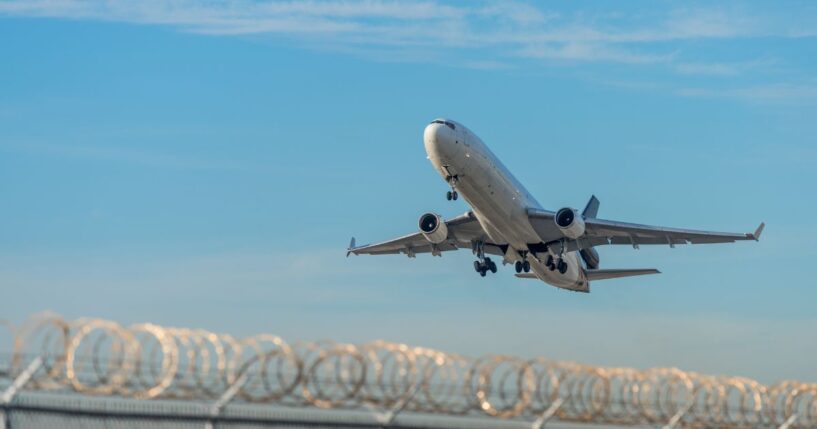 A deportation plane taking off over a barbed wire fence at Miami International Airport.