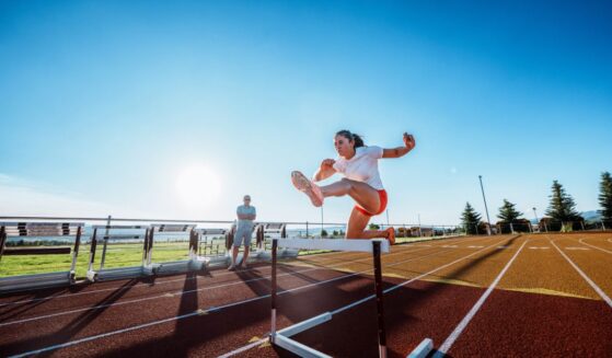 A female collegiate athlete powers over a hurdle at full speed during an intense solo training session with her coach observing nearby.
