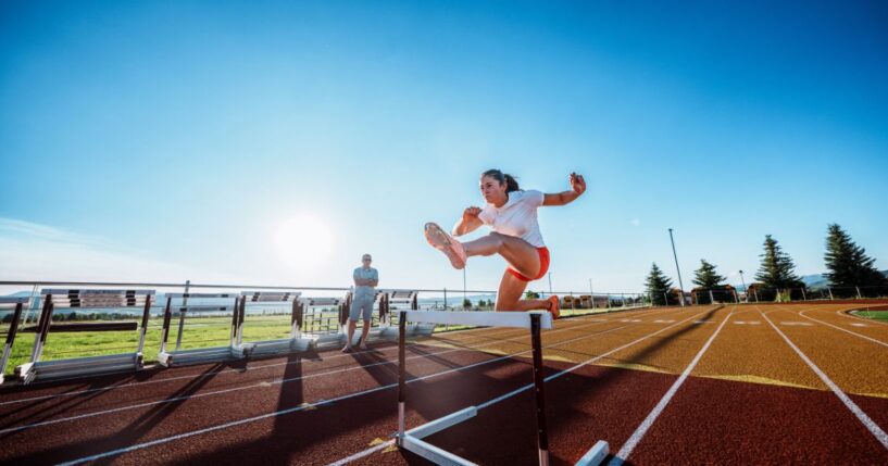A female collegiate athlete powers over a hurdle at full speed during an intense solo training session with her coach observing nearby.