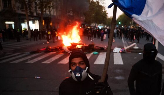 A protestor with a gas mask holds a French flag in front of a fire at the Boulevard des Capucines during a demonstration in Paris on Oct. 17, 2026.
