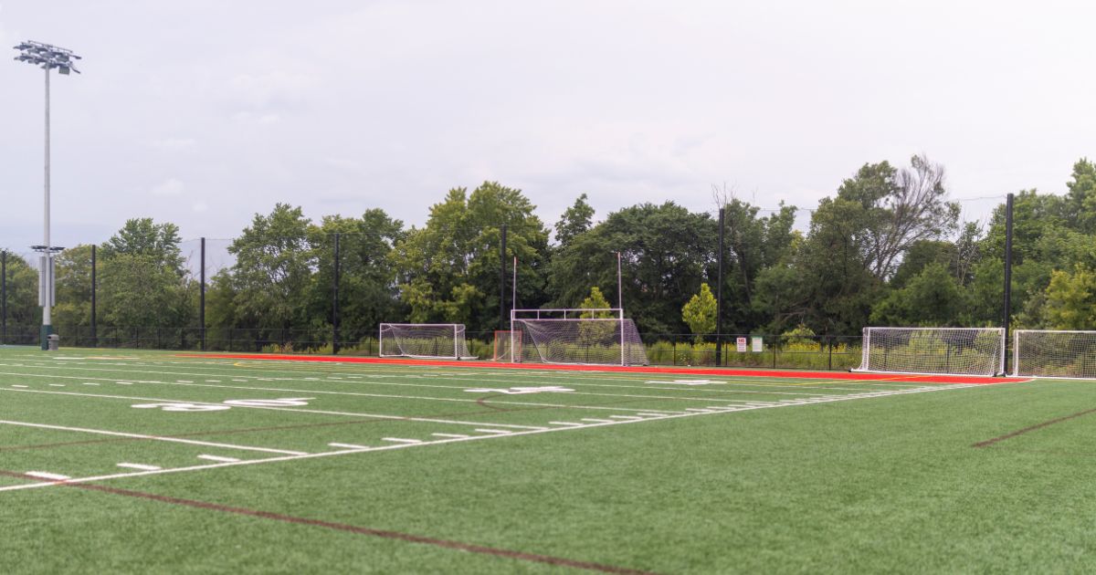 An artificial turf athletic field featuring yard lines, multiple goalposts, a red running track, and a towering stadium light pole.