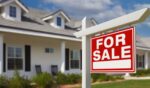 A For Sale sign stands in front of a house planted in the front yard.