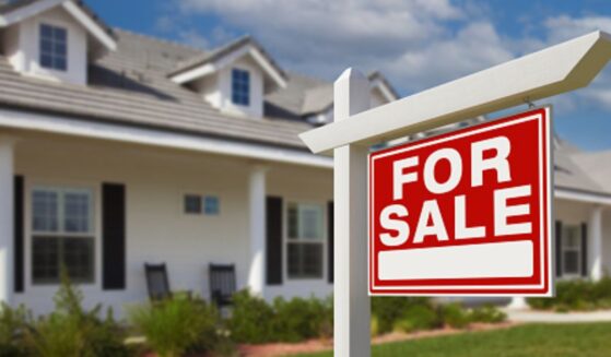 A For Sale sign stands in front of a house planted in the front yard.