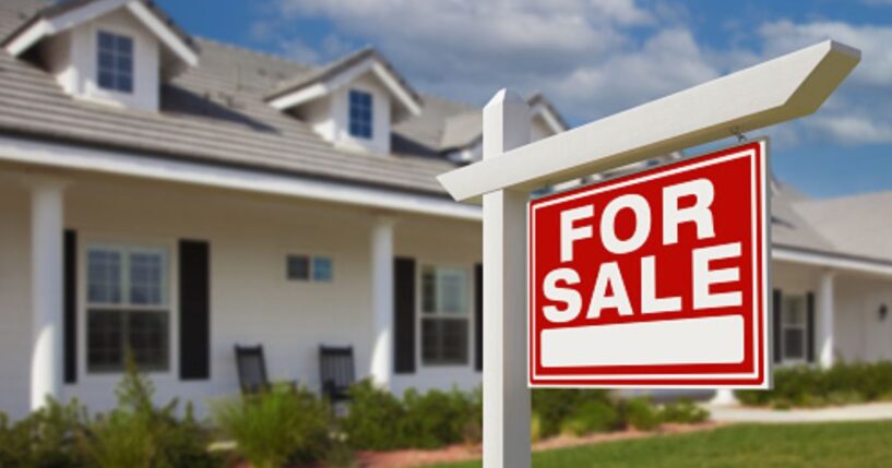 A For Sale sign stands in front of a house planted in the front yard.